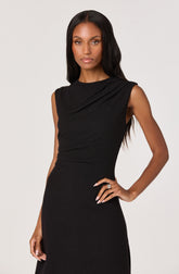 A woman is wearing a sleeveless black dress with a high neckline and subtle pleats on the shoulders. The dress is fitted at the waist and has a smooth, elegant silhouette. She has long, wavy black hair and is accessorizing with rings on her fingers.